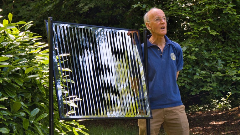 The artist Michael Joseph standing by his artwork at Claremont. The artwork is a large, rectangular, reflective panel with vertical stripes.
